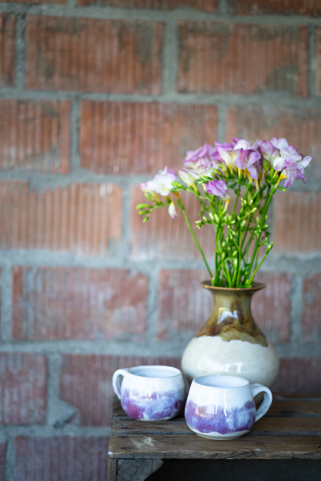 Handgetöpferte Vase mit Freesien vor einer Backsteinwand, davor zwei Becher